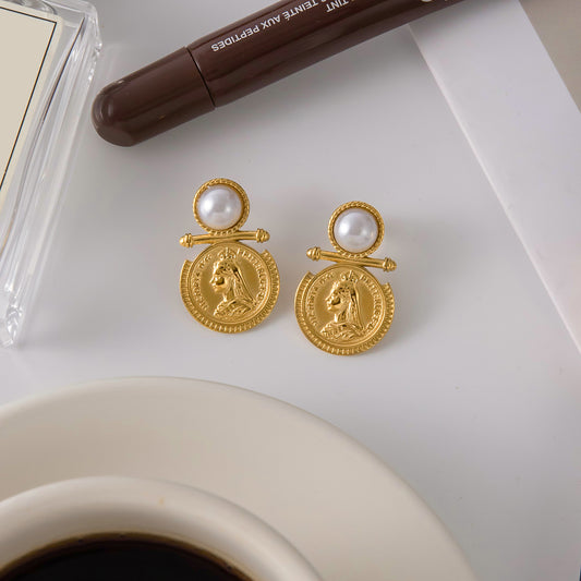 Aura Coin Pearl Earrings displayed on a table next to a coffee cup, featuring gold medallions and pearls.
