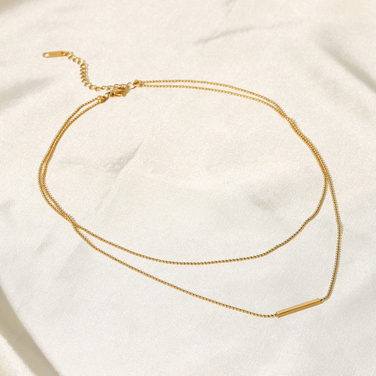 Layered bar necklace on a soft fabric background, featuring two gold-plated chains and a sleek horizontal bar pendant.