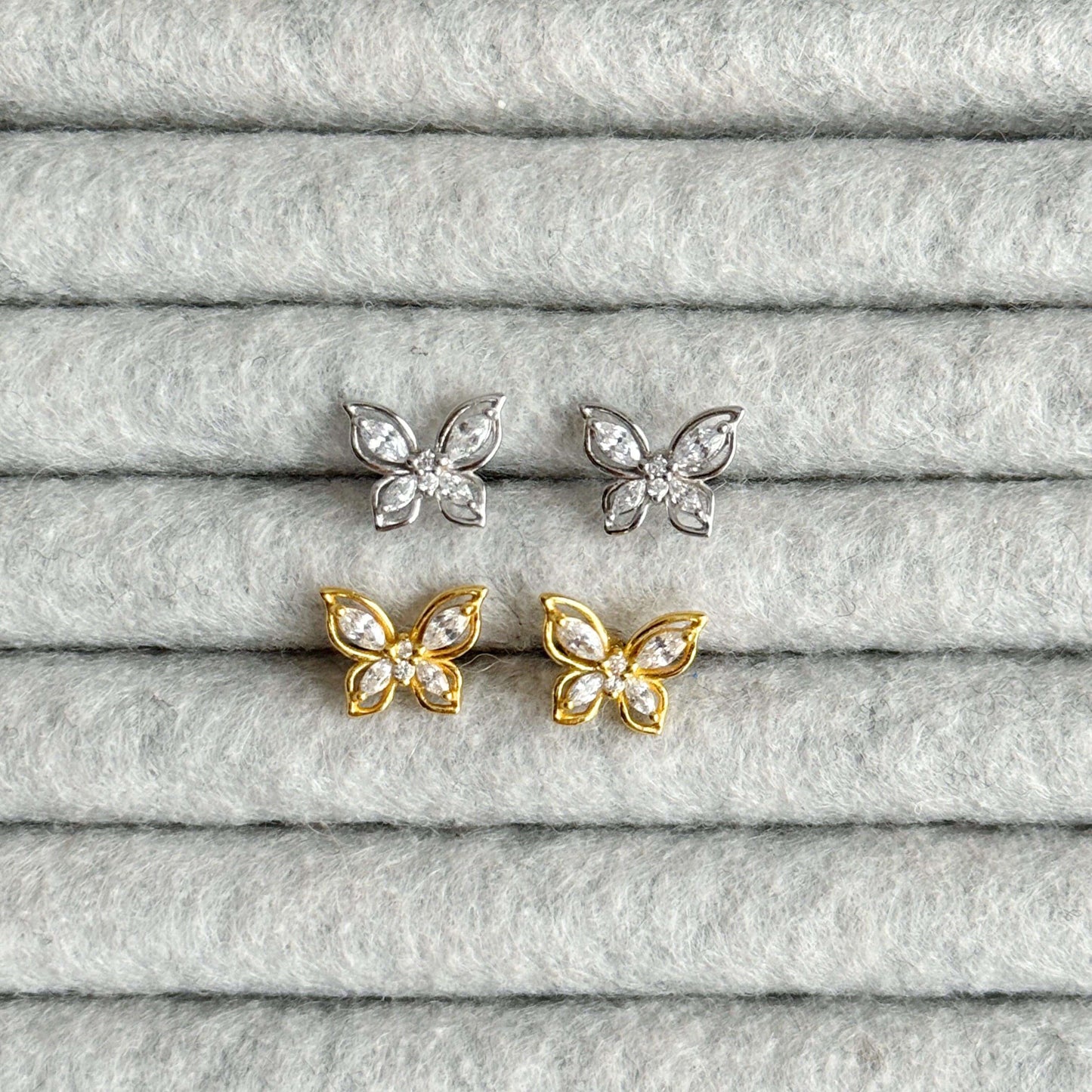 Elegant butterfly stud earrings in silver and gold, featuring shimmering zirconia, displayed on soft gray fabric.