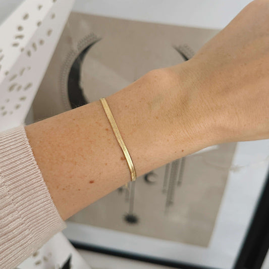 Close-up of a wrist wearing a gold snake bracelet, showcasing its delicate herringbone design against a stylish background.