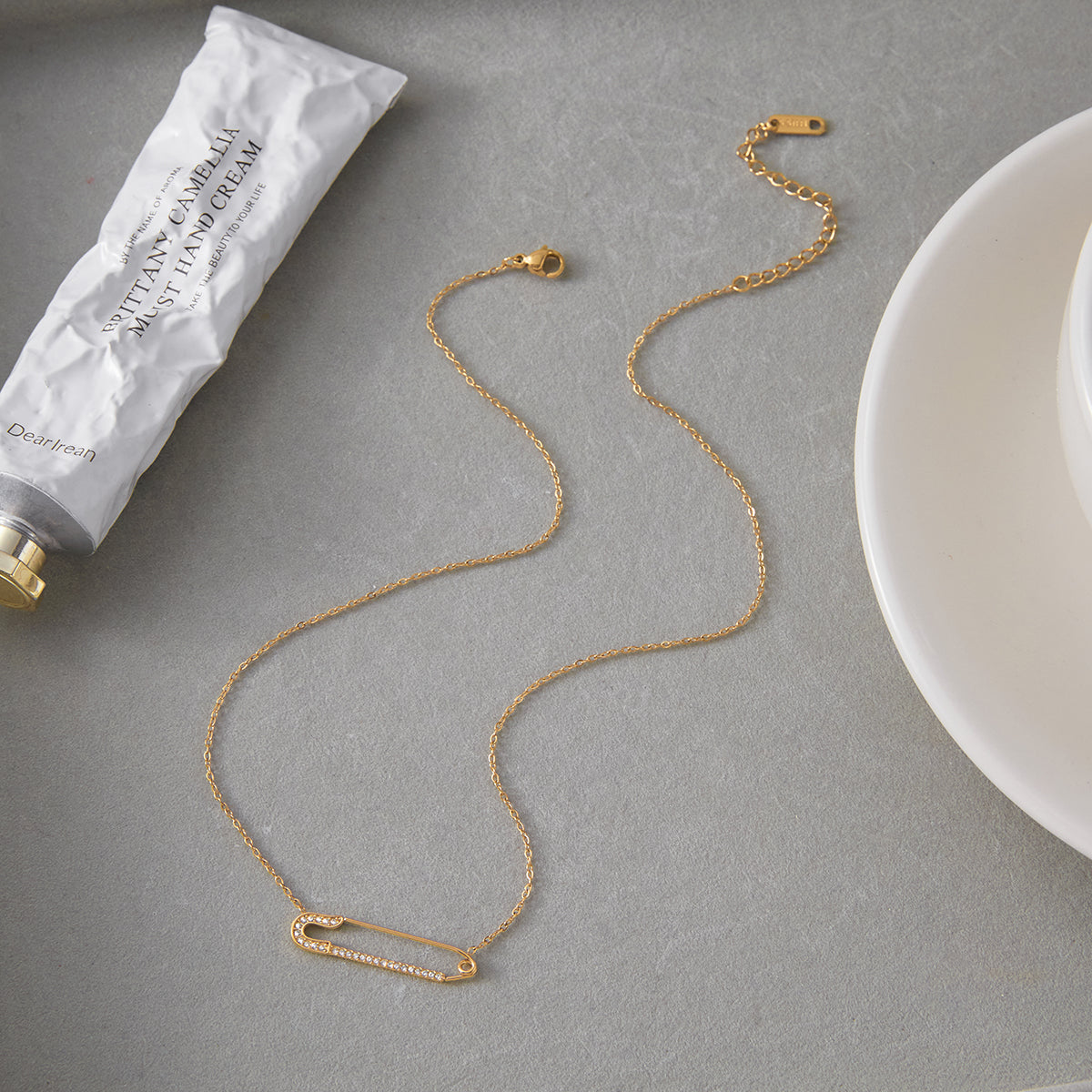 Safety Pin Necklace | Waterproof & Tarnish Free displayed on a grey surface with hand cream and a plate.