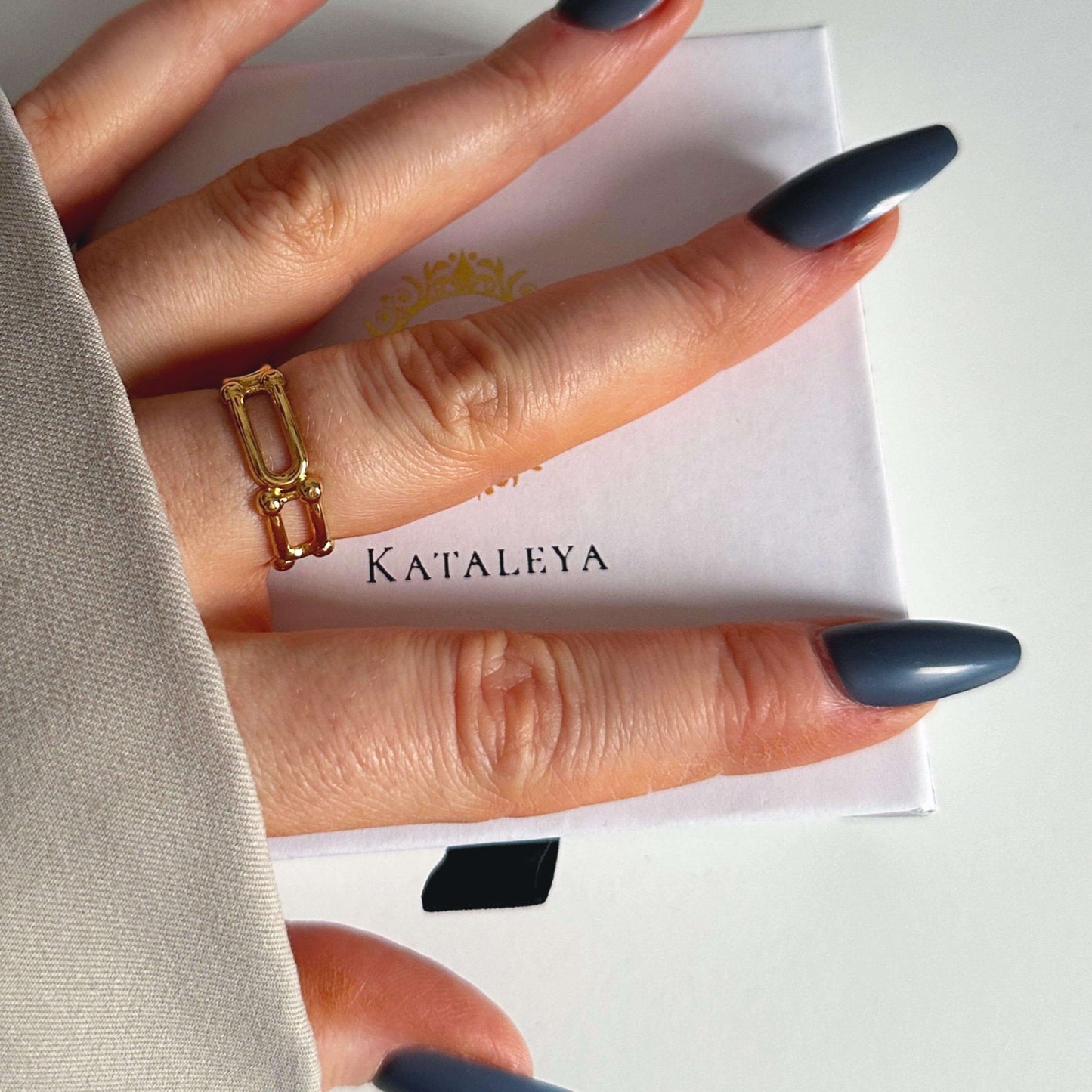 Close-up of a hand wearing a stylish gold U-ring with long, manicured nails on a white KatAleya box.