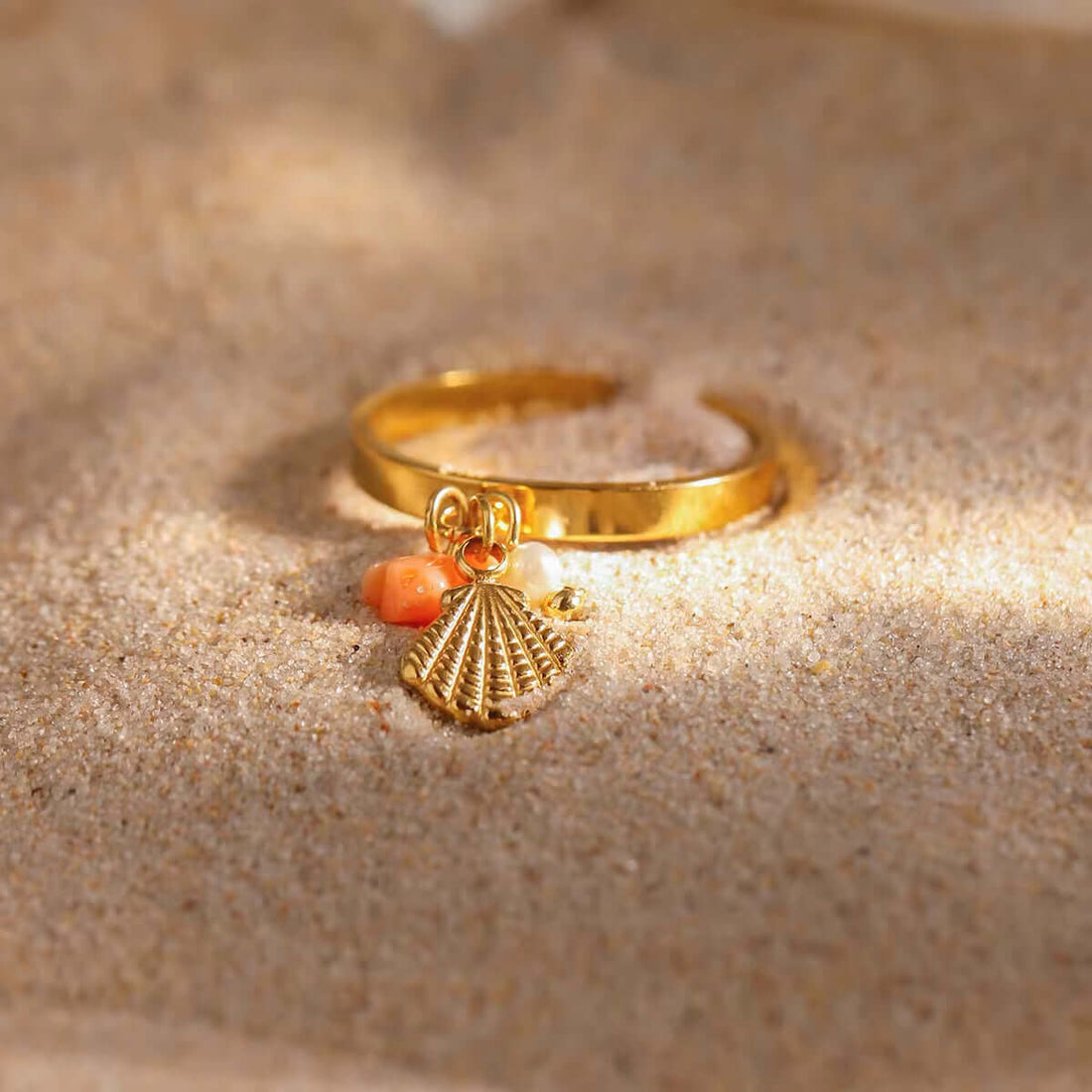 Jewelry Inspired by the Ocean: A golden ring with a seashell charm and coral bead resting on sandy beach.