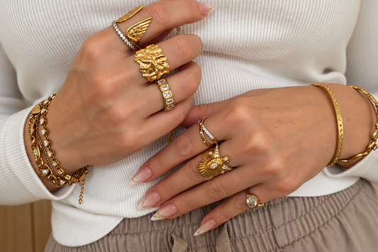 Hands wearing stacked gold rings in chunky, pavé and signet styles, with gold bracelets, against a white knitted jumper.
