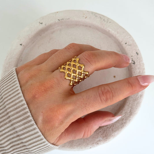 A hand wearing a gold mesh open ring with a lattice design, styled elegantly on a white plate.
