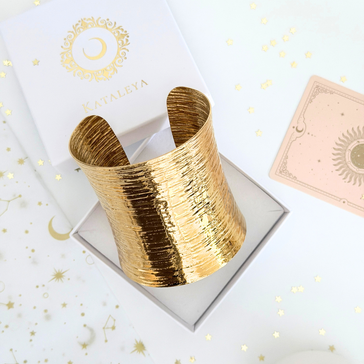 Theia Wide Cuff displayed in a box, showcasing its bold golden design with textured surface against a decorated background.