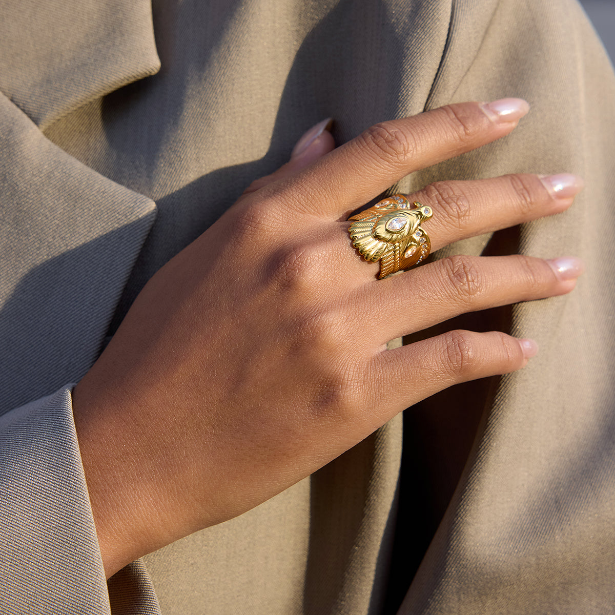 Hand wearing a golden Nekhbet eagle statement ring on a beige outfit, showcasing its bold design and elegance.