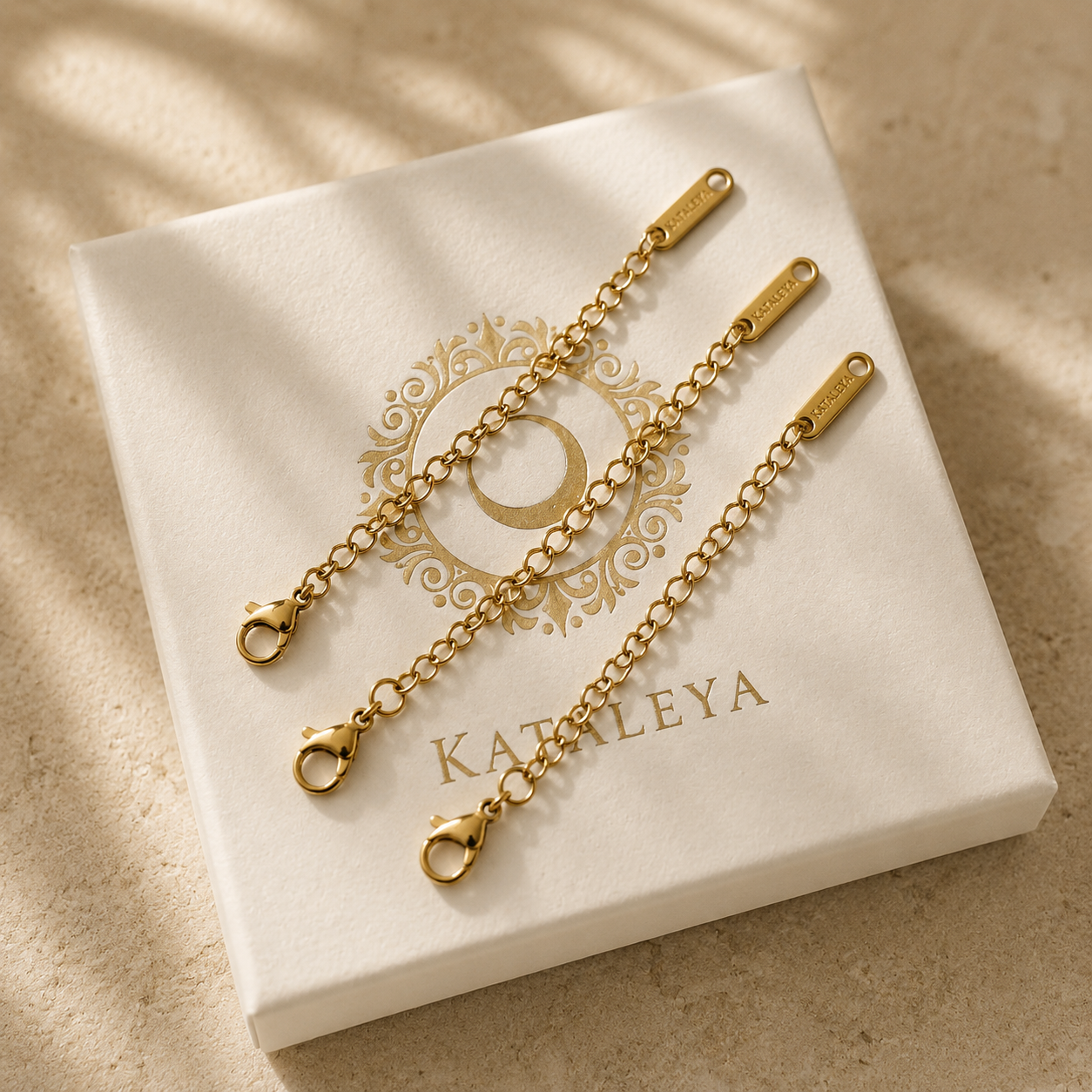 Two gold-tone chain extender links with lobster clasp ends, displayed on a white KATALEYA jewelry box on a beige surface with soft leaf shadows.