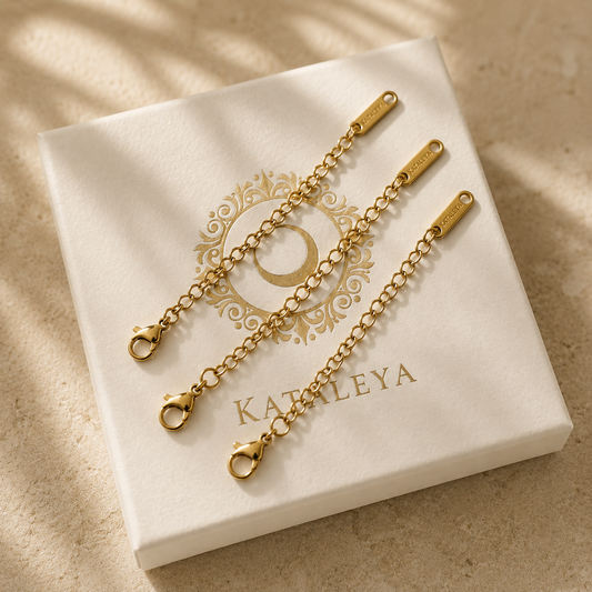 Two gold-tone chain extender links with lobster clasp ends, displayed on a white KATALEYA jewelry box on a beige surface with soft leaf shadows.