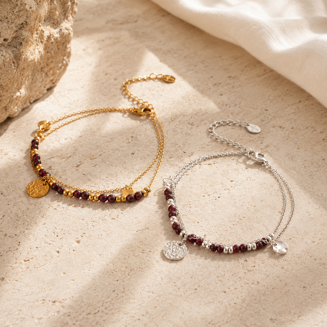 Two delicate beaded bracelets displayed on a textured beige stone surface. One bracelet is gold-toned with dark red and metallic beads, featuring a round gold charm.